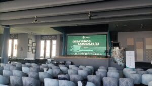 Sala de conferencias con filas de sillas vacías frente a una pantalla grande que muestra detalles del evento "Desayunos Laborales '23".
