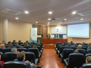 Público sentado en una sala de conferencias viendo una presentación de dos oradores en un podio, con una pantalla de proyección que muestra el contenido.