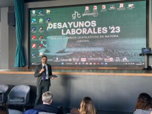 Una persona habla en el escenario durante el evento "Desayunos Laborales '23". Una gran pantalla muestra detalles del evento y logotipos en el fondo. Los miembros del público están sentados en primer plano.