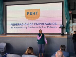 Una mujer habla por un micrófono frente a una gran pantalla que muestra el logo de la Federación de Empresarios de Hostelería y Turismo de Las Palmas. Los miembros del público están sentados al frente.