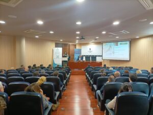 Una sala de conferencias con un público sentado en filas frente a un escenario. Hay tres presentadores en una mesa y un orador en un podio. Se muestra una presentación en la pantalla.