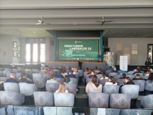 Un grupo de personas sentadas en una sala frente a un escenario con una pantalla grande que muestra "Desayunos Laborales '23.