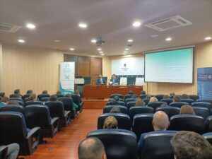 Se está dando una conferencia en una sala de conferencias con dos presentadores en un podio y un público sentado en filas. Se muestra una presentación en una pantalla.