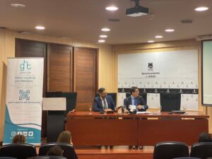 Dos hombres de traje se sientan en una mesa redonda en una sala de conferencias. A la izquierda hay una pancarta y en la pared hay un logotipo del Ayuntamiento de Adeje. Los asistentes se sientan en primer plano.