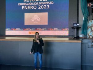 Una mujer se para frente a una pantalla grande que muestra una presentación titulada "Novedades Protección por Desempleo Enero 2023.