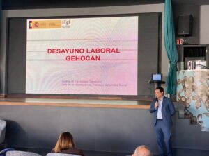 Un hombre de traje habla a través de un micrófono junto a una pantalla de presentación que muestra una diapositiva con el texto titulado “Desayuno Laboral” en un contexto de conferencia.