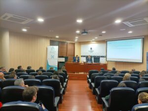 Las personas en una sala de conferencias se sientan frente a una pantalla de presentación y a los oradores en el podio delantero.