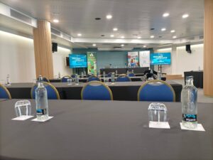 Sala de conferencias con filas de mesas y sillas, botellas de agua y vasos en las mesas y pantallas de presentación en la parte delantera.