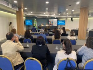 Público sentado en una sala de conferencias, frente a un panel de oradores que discuten una presentación.