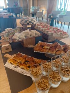 Una mesa de buffet con croissants, sándwiches, parfaits de yogur y postres, ubicada en un espacioso comedor con sillas y mesas al fondo.