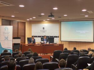 Dos oradores se sientan en un podio y se dirigen a un público en una sala de conferencias. En una pantalla se proyecta una presentación titulada "Novedades legislativas y judiciales".