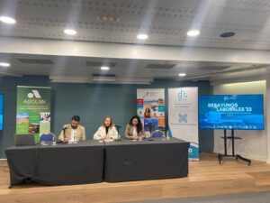 Tres personas están sentadas en una mesa de conferencias con micrófonos. Detrás de ellas hay pancartas y una pantalla que muestra "Desayunos Laborales '23".