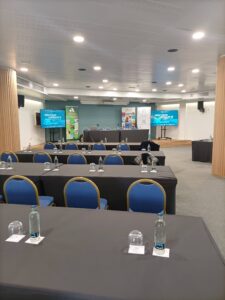 Sala de conferencias con filas de mesas y sillas, cada mesa con botellas de agua y vasos. Se ven dos pantallas grandes al frente.