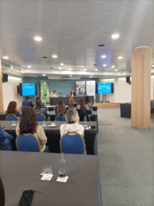 Una sala de conferencias con personas sentadas frente a una pantalla de presentación. Los presentadores están al frente y hay carteles informativos cerca.