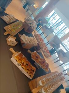 Mesa de buffet con distintos bocadillos, pasteles y parfaits. Sala luminosa con grandes ventanales y sillas al fondo.