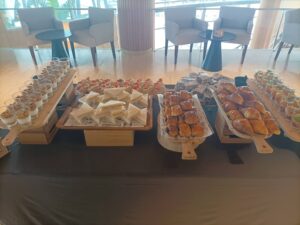 Mesa de buffet con una variedad de bocadillos, que incluyen sándwiches, pasteles, parfaits de yogur y copas de fruta, ubicada en una moderna zona de estar.