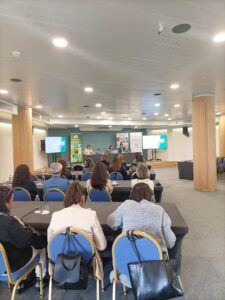Personas que asisten a una conferencia en una sala con tres presentadores al frente. Los asistentes están sentados en mesas con manteles negros, frente a pantallas que muestran diapositivas de presentaciones.
