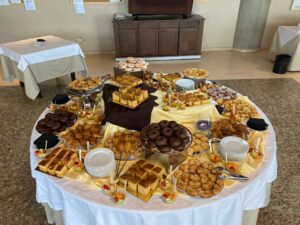 Una mesa redonda está llena de diversos pasteles, incluidos donuts, pasteles y muffins, todos expuestos sobre un mantel blanco.