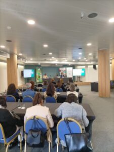 Una sala de conferencias con personas sentadas en mesas frente a un panel de oradores. Se ven pantallas de presentación y se muestran varios carteles en el frente.