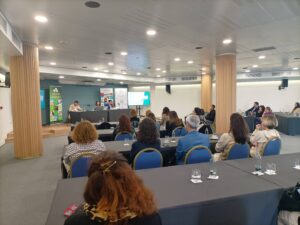 Una sala de conferencias con los asistentes sentados frente a un panel de oradores en una mesa. El equipo de presentación se ve al fondo.
