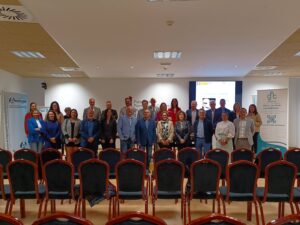 Un grupo de personas vestidas formalmente se encuentra en una sala de conferencias frente a una pantalla de presentación y sillas vacías.