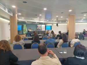 Un grupo de personas se sienta en una sala de conferencias, frente a un panel de oradores con pantallas de presentación a ambos lados.