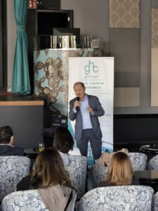 Un orador con traje se dirige a un público en un espacio cerrado, de pie frente a una pancarta con el texto "GEHOCAN" y "EL VALOR DE LAS PERSONAS".