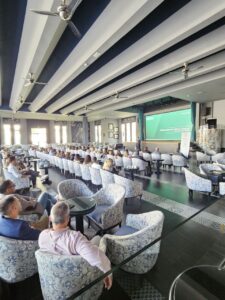 Una sala de conferencias con los asistentes sentados frente a una pantalla grande que muestra una presentación. La sala tiene sillas estampadas y ventiladores de techo.