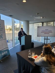 Persona hablando en un podio en una sala de conferencias con vista a una ciudad a través de grandes ventanales. Los asistentes están sentados en primer plano. Una pancarta y una pantalla de proyección son visibles al lado del orador.