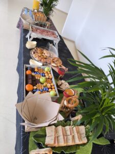 Una mesa buffet con sándwiches, pasteles, muffins, brownies, frutas, jugos y plantas como decoración.