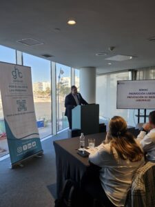 Un orador realiza una presentación en un podio en una sala de conferencias. Una pancarta y una pantalla muestran información sobre prevención de riesgos laborales. Los asistentes se sientan y escuchan.