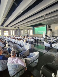 Un grupo de personas sentadas en una sala de conferencias con sillas estampadas, frente a una pantalla que muestra una presentación.