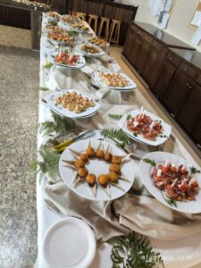 Una mesa de buffet con diversos aperitivos en platos blancos, entre ellos brochetas, pasteles y rollitos de carne, está decorada con helechos.