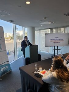 Un hombre da una presentación sobre prevención de riesgos laborales en una sala de conferencias, con una pantalla que muestra el tema. Los miembros de la audiencia observan atentamente.