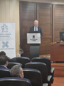 Un hombre de traje habla en un podio en una sala revestida de madera. Los miembros del público están sentados y se ve una pancarta a la izquierda.