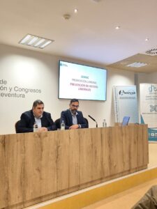 Dos hombres sentados en una mesa durante una presentación titulada "SEMAC Promoción Laboral Prevención de Riesgos Laborales" en una sala de conferencias.