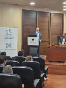 Un hombre se encuentra de pie en un podio hablando con los asistentes sentados en una sala de conferencias. A su lado se ve una pancarta con texto y logotipos.