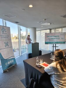 Una mujer se encuentra en un podio y realiza una presentación ante los asistentes a un evento empresarial. Una pancarta y una pantalla muestran el lema del evento: "Desayunos Laborales '24".