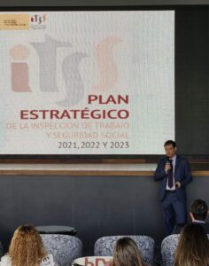 Un hombre habla en un escenario junto a una diapositiva proyectada titulada "Plan Estratégico de la Inspección de Trabajo y Seguridad Social 2021, 2022 y 2023". Los miembros del público están sentados en primer plano.