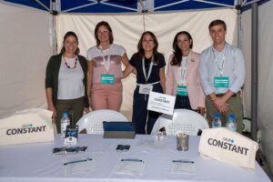 Cinco personas están de pie detrás de una mesa de exposición con el logo de "Grup Constant" y sonríen a la cámara. En la mesa se exhiben folletos y bolsas de tela con el logo. Están bajo una carpa azul y blanca.