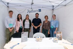 Seis personas de pie en un stand de Radisson Blu bajo una carpa blanca, sonriendo a la cámara. Frente a ellas hay una mesa con folletos y bolsas de tela.