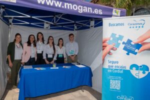 Un grupo de seis personas se encuentra detrás de una mesa en un puesto, con una pancarta que promociona "Cordial". La pancarta presenta piezas de rompecabezas y el texto "Seguro que encajas en Cordial".