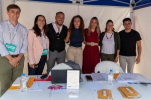 Siete personas de pie juntas detrás de una mesa con papeles, carpetas y botellas de agua, bajo un dosel.