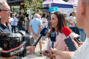Una mujer habla a través de varios micrófonos sostenidos por periodistas en un entorno al aire libre con gente de fondo.