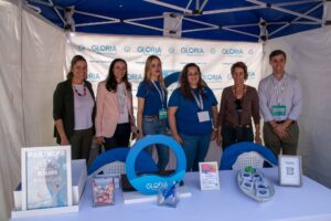 Siete personas se encuentran de pie bajo una carpa azul y blanca con la marca "Gloria". Están ubicadas detrás de una mesa que exhibe artículos promocionales.