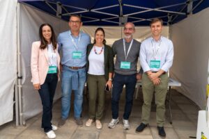 Cinco personas están juntas bajo una carpa, todas sonriendo a la cámara. Llevan ropa informal y llevan cordones con etiquetas con sus nombres.