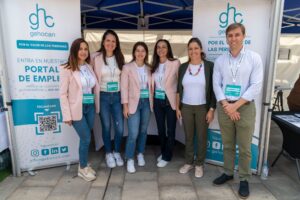 Siete personas están de pie sonriendo frente a un stand con carteles "gehocan", portando etiquetas con sus nombres y vestimenta informal de negocios.