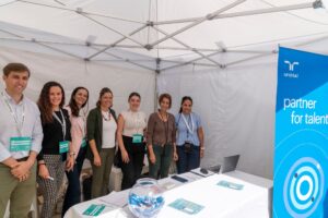 Siete personas de pie en una carpa con una pancarta azul que dice "Acompañamiento para el talento", una mesa con folletos y un bol de caramelos. Sonríen y llevan etiquetas con sus nombres.