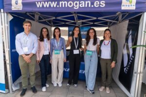Seis personas están de pie sonriendo bajo un dosel azul con el sitio web "www.mogan.es" sobre él. Llevan ropa de trabajo informal y llevan etiquetas con sus nombres.