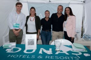 Cinco personas de pie detrás de una mesa con una pancarta de Dunas Hotels & Resorts, bajo una carpa blanca. Sobre la mesa hay un cartel con un código QR y papeles dispersos.